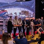 A group of eight men and two women stand on a red carpet in front of a large screen displaying an airplane and palm trees. They are each holding a pair of scissors and cutting a long orange ribbon. The seven audience members in front of them are photographing them with cell phones.