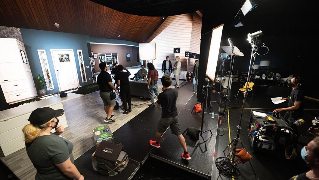 Crew members set up cameras and make adjustments to a virtual living room backdrop between scenes.